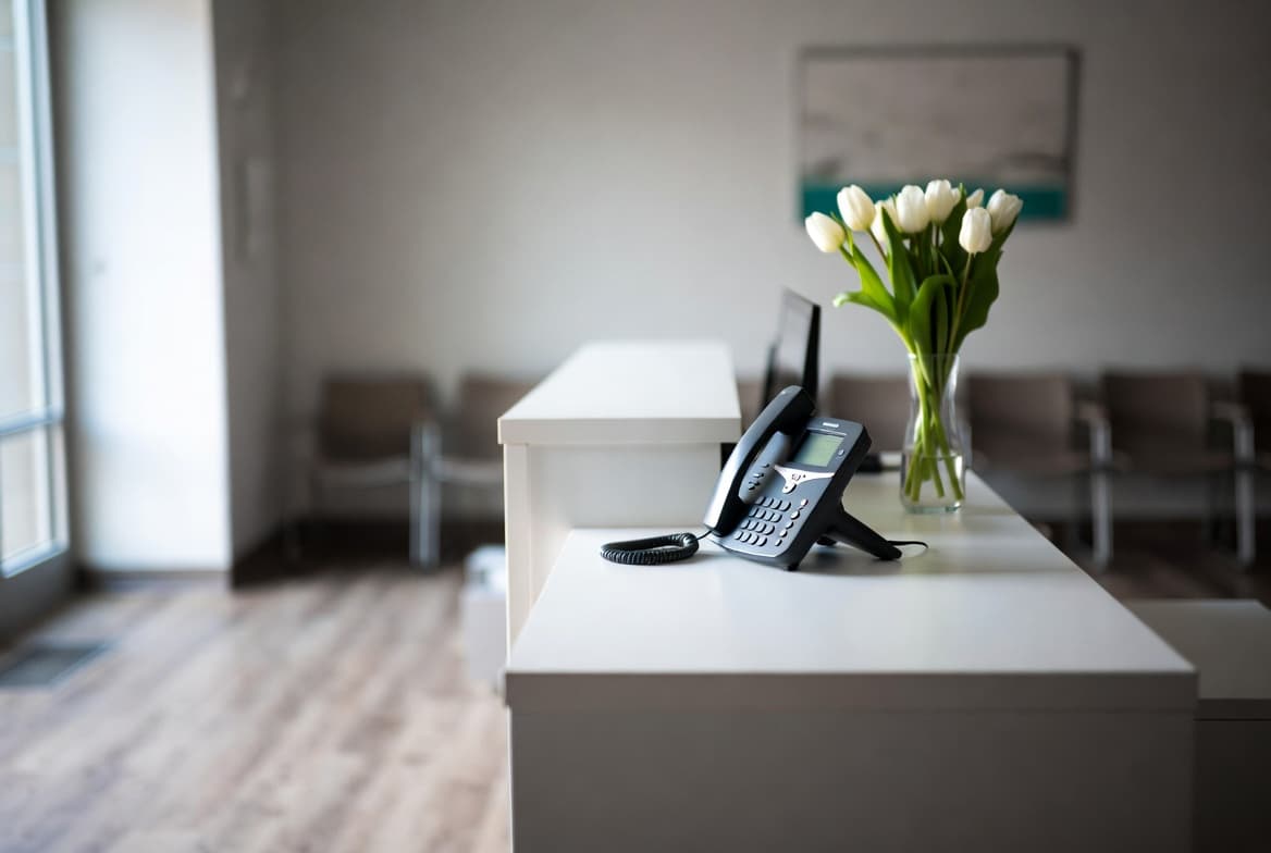 An empty school front office at dawn — a single phone on a white reception desk, quiet, before the day begins.